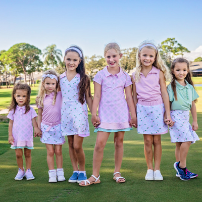 Six young girls ranging from toddler to junior stand on a golf course modeling pink, white, and green girls golf outfits.
