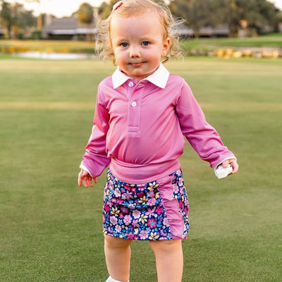 A toddler girl stands on a golf course modeling a long sleeve pink polo and a baby skort.