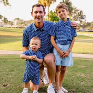 two young boys and a grandfather stand on a golf course modeling matching family outfits consisting of blue striped polo shirts and blue chino shorts