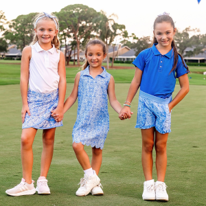 Five young girls jumping on a golf course modeling pink and blue golf outfits consisting of girls dresses, girls polo shirts, and skorts for girls