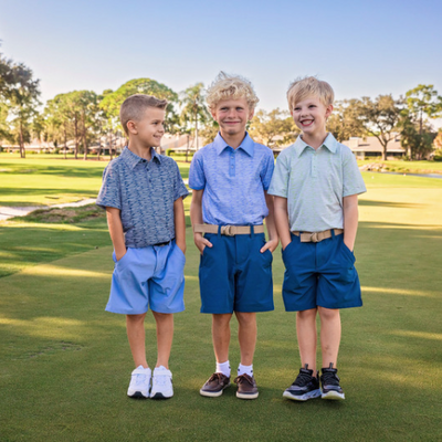 Three young boys stand on a golf course modeling blue golf shorts and blue and green performance polos