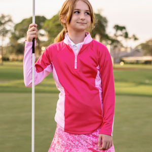 A young girl stands on a golf course modeling a pink quarter zip jacket.