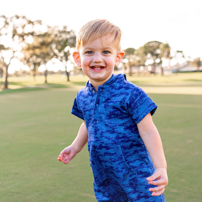 Young boy in a blue Turtles and Tees romper standing on a grassy field with trees in the background.