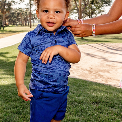 Toddler boy wearing a Turtles and Tees blue textured polo shirt and blue shorts standing outdoors with a person's hand on their shoulder.