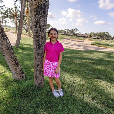Young girl in Turtles and Tees pink polo shirt and skirt standing next to a tree on a golf course