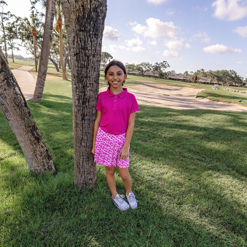 Young girl in a Turtles and Tees pink polo shirt and skirt standing next to a tree on a golf course.