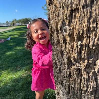 Toddler girl in a Turtles and Tees pink golf dress peeking from behind a tree