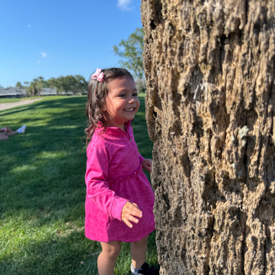 Toddler in a Turtles and Tees pink golf dress standing behind a tree smiling.