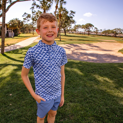 Young boy wearing a Turtles and Tees blue wave patterned golf polo and shorts standing on grass with trees in the background