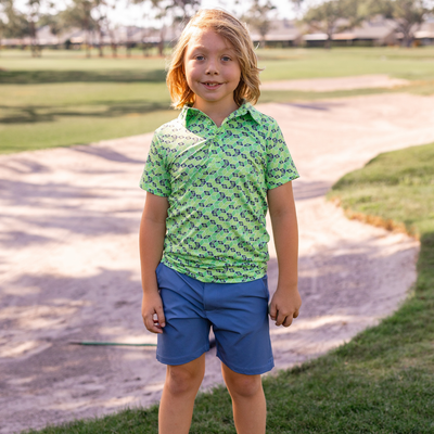 Child wearing a Turtles and Tees green patterned golf shirt and blue shorts standing on a golf course.