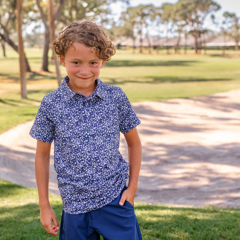 Young boy wearing a Turtles and Tees blue floral patterned polo shirt and navy shorts standing in a golf course.
