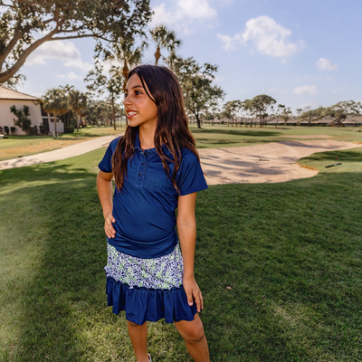 Young girl in a Turtles and Tees blue polo shirt and blue floral golf skort standing on a grassy area with trees and a building in the background.