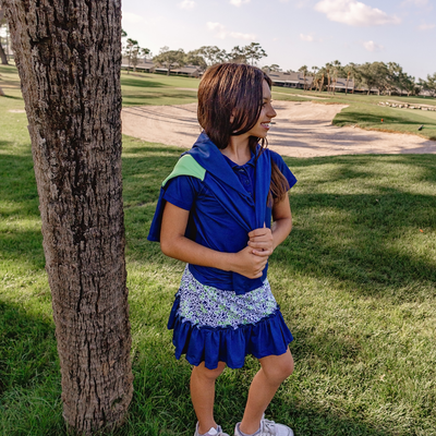 Girl in a Turtles and Tees blue polo shirt and blue floral golf skort standing on a golf course with a tree in the foreground
