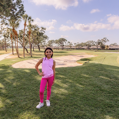 Young girl in Turtles and Tees pink golf outfit standing on a golf course with trees and sky in the background