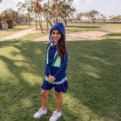 Girl in a blue and green Turtles and Tees golf jacket and skort, standing on a grassy area with trees in the background