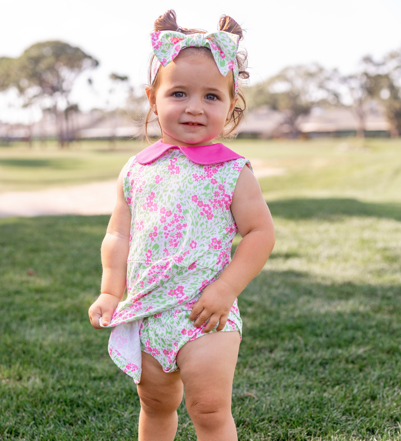 A toddler girl modeling a Turtles and Tees floral print pink and green infant and toddler romper and a floral bow headband