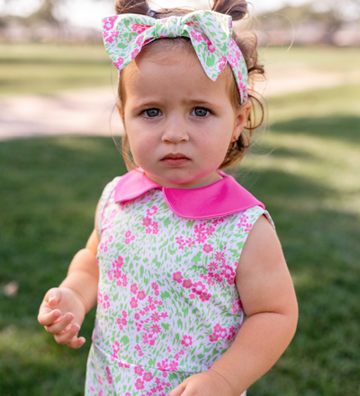 A toddler girl modeling a Turtles and Tees floral print pink and green infant and toddler romper and a floral bow headband