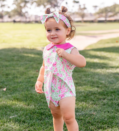 A toddler girl modeling a Turtles and Tees floral print pink and green infant and toddler romper and a floral bow headband