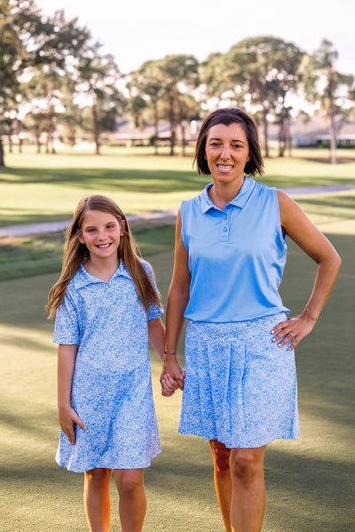 A woman stands on a golf course modeling a blue floral golf skort and blue sleeveless polo, next to a child wearing a matching blue floral dress