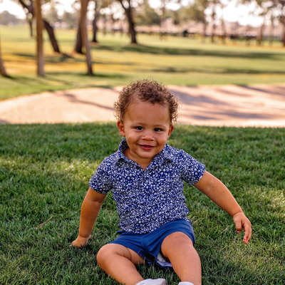 Toddler boy sitting on grass in a park wearing a Turtles and Tees blue floral polo shirt and navy shorts.