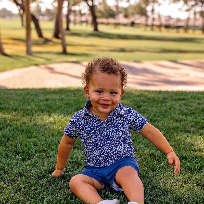 Toddler boy sitting on grass in a park wearing a Turtles and Tees blue floral polo shirt and navy shorts.