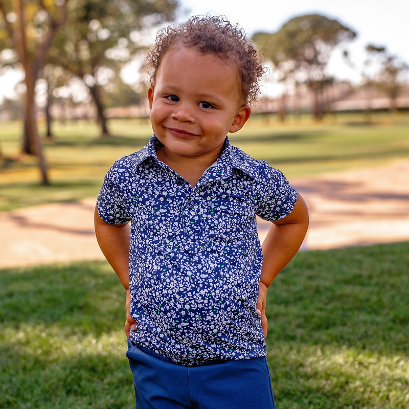 Toddler boy wearing a Turtles and Tees blue floral polo and blue shorts standing in a park.