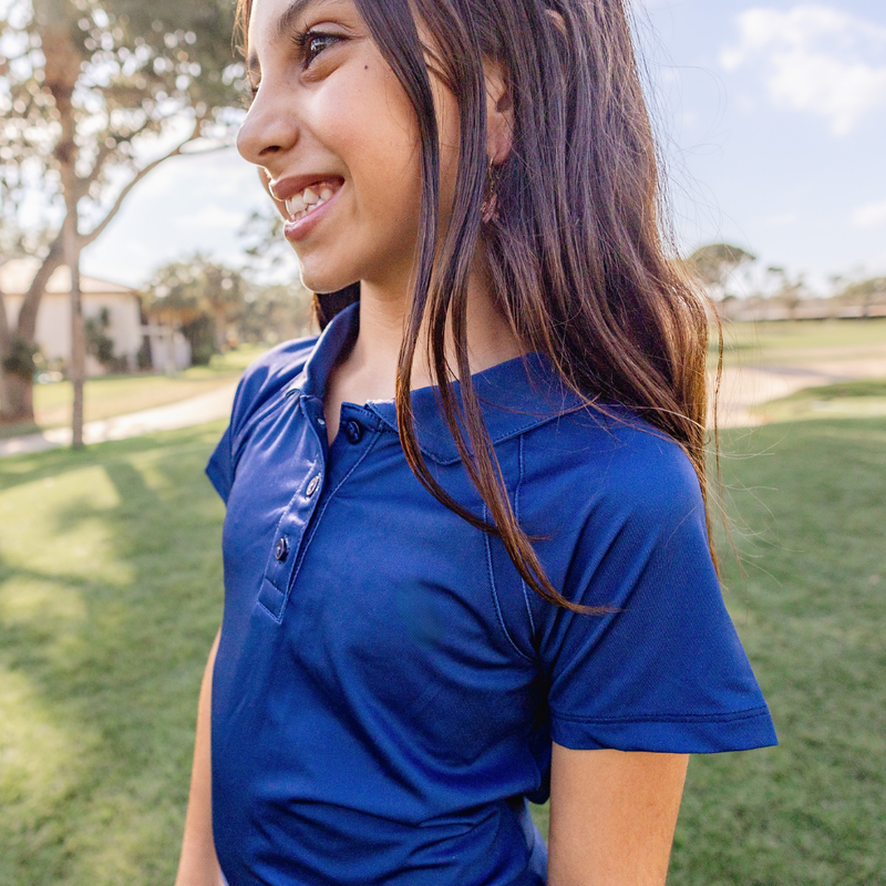A young girls standing on a golf course wearing a Turtles and Tees navy blue cap sleeve polo.
