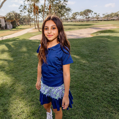 Young girl in a Turtles and Tees blue top and floral skirt standing on grass with trees and a path in the background