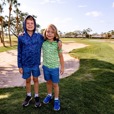 Two children standing on a golf course wearing Turtles and Tees blue and green golf apparel with palm trees in the background