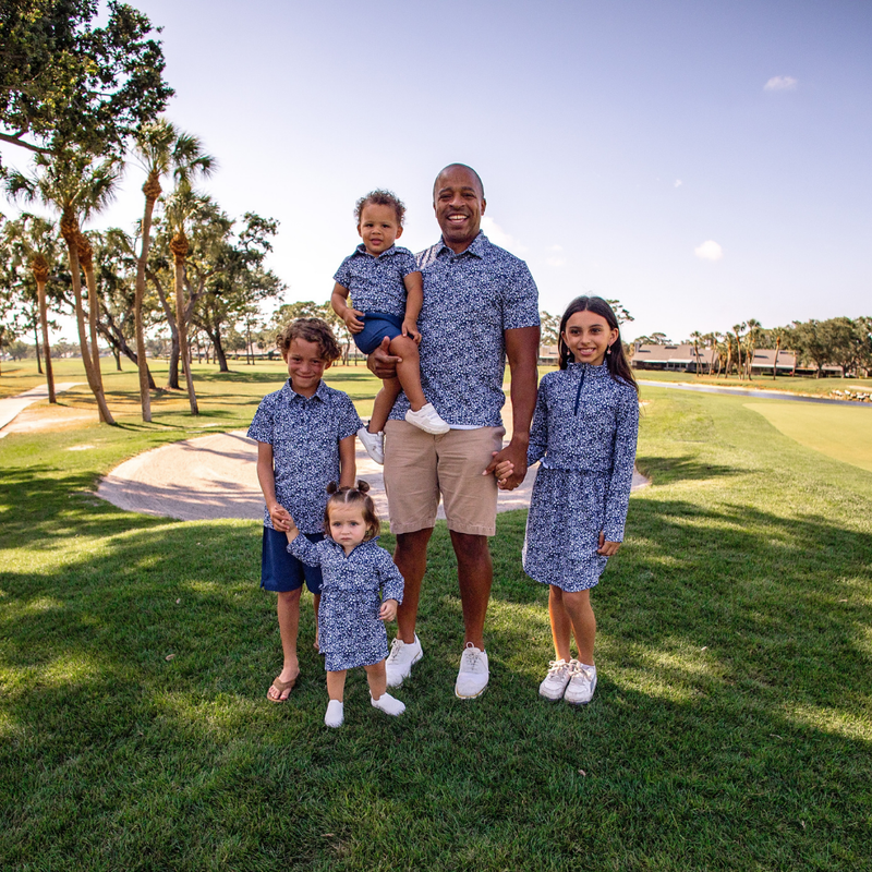 Family of five posing in Turtles and Tees matching blue floral outfits on a golf course with trees and clear sky in the background