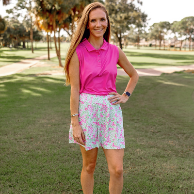 A woman stands on a golf course modeling a pink and green floral skort and pink sleeveless polo