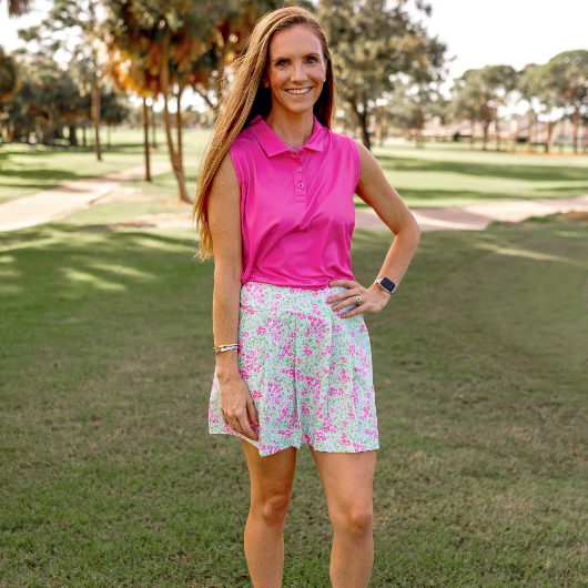 A woman stands on a golf course modeling a pink and green floral skort and pink sleeveless polo