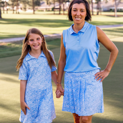 A woman and girls stand on a golf course modeling Turtles and Tees floral blue golf skort, floral blue golf dress, and blue sleeveless polo