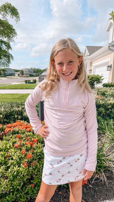 Young blonde girl stands by shrubbery wearing a pink quarter zip and floral golf skort