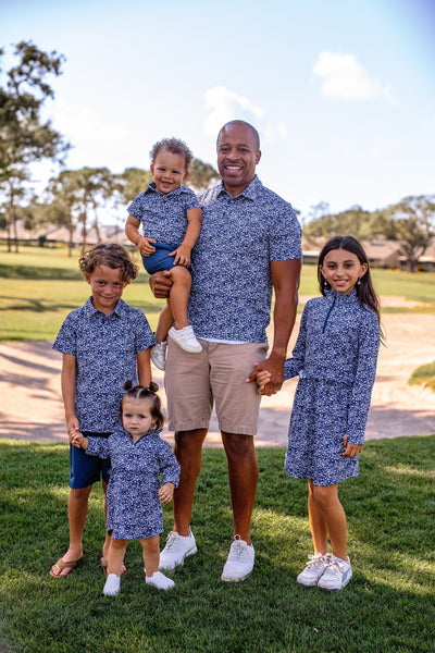 Family of five wearing Turtles and Tees matching blue floral outfits standing on grass with trees and a clear sky in the background.