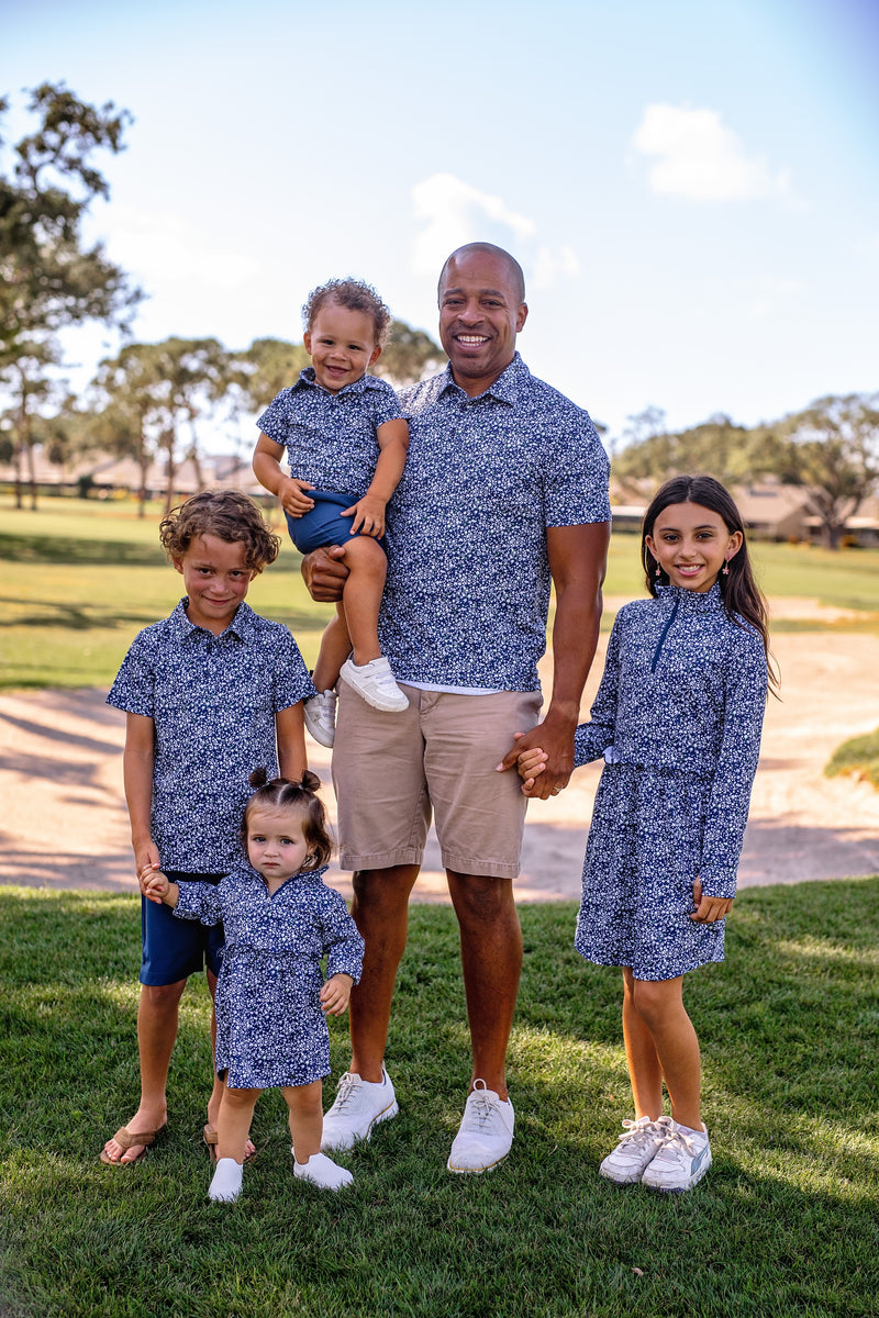 Family of five wearing Turtles and Tees matching blue floral outfits standing on grass with trees and a clear sky in the background.