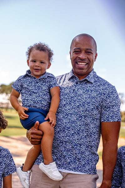 Man holding a child in Turtles and Tees matching blue floral outfits with a clear sky background