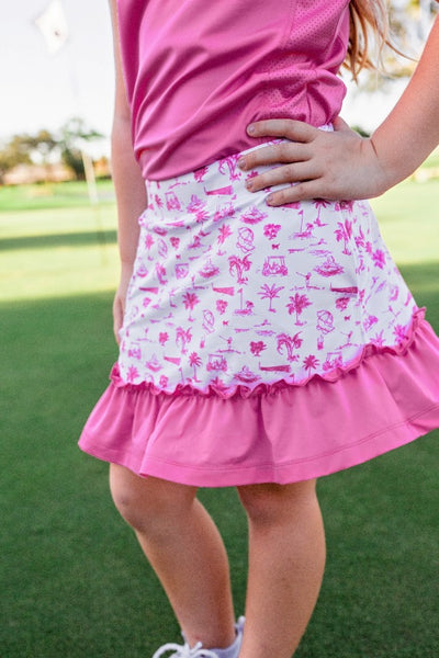 A young girl standing on a golf course modeling an outfit that consists of a hot pink polo paired with a golf skort. The skort is white with beach and golf pink prints on it, and has a pink ruffled hem.