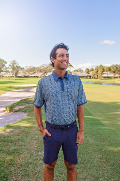 A man stands on a golf course modeling navy blue chino shorts and a navy blue and green performance polo shirt.