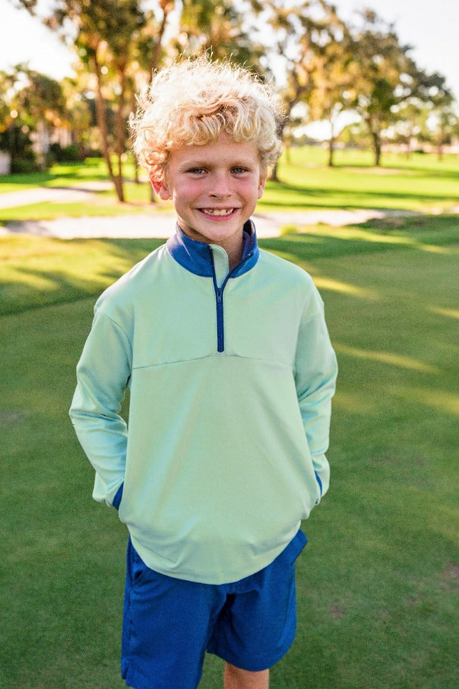 A young boy stands on a golf course modeling a light green quarter zip with a blue zipper and navy blue chino shorts