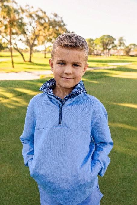 A young boy stands on a golf course modeling a blue quarter zip sweatshirt