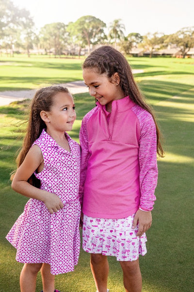 Two young girls stand outside, about to hug. One is wearing a pink and white printed sleeveless dress, and the other is wearing a pink pullover and pink and white golf skort.