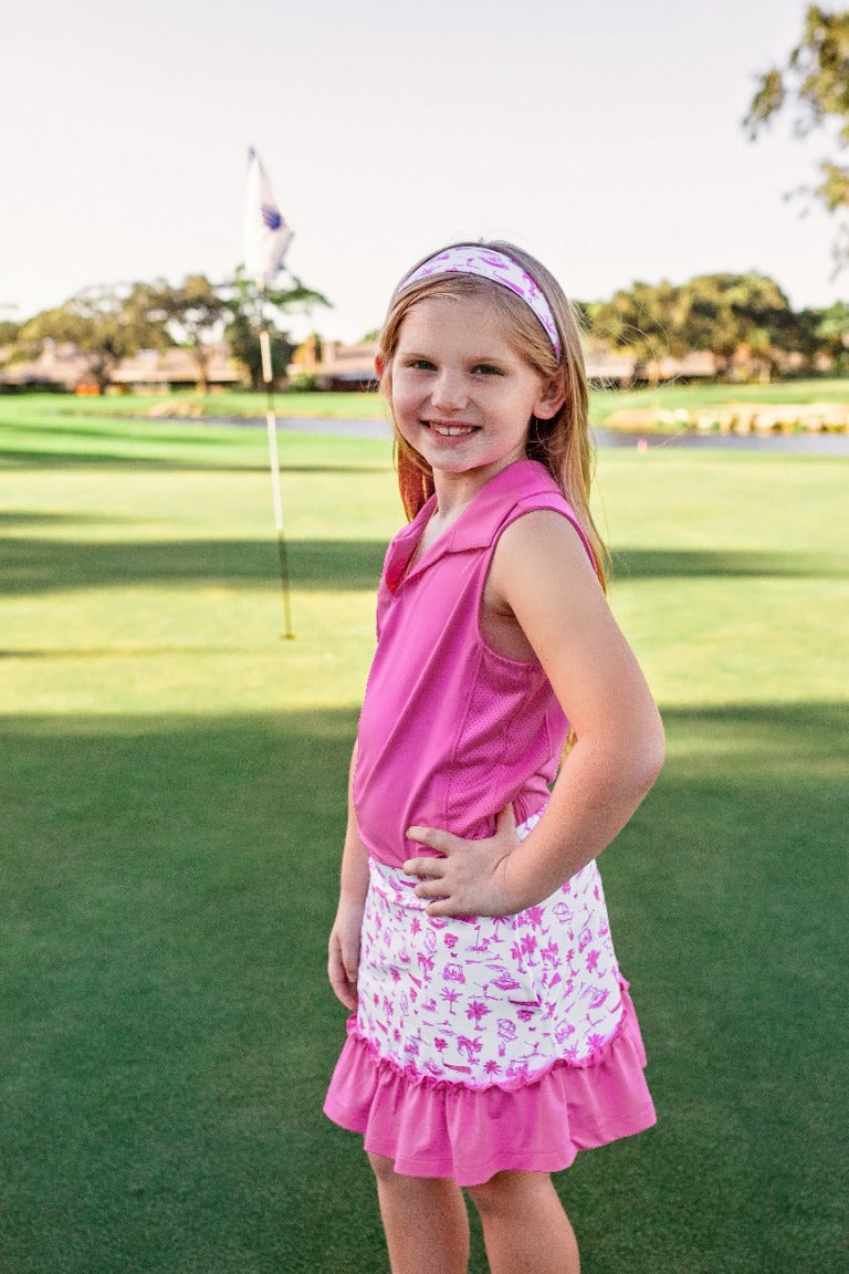 A young girl stands on a golf course modeling a pink sleeveless polo shirt and white and pink skort.