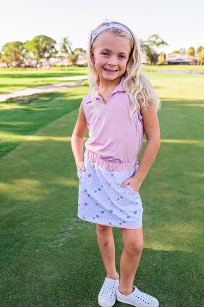A young girl stands on a golf course modeling a pink or light purple sleeveless polo shirt and a pink floral multicolored skort.