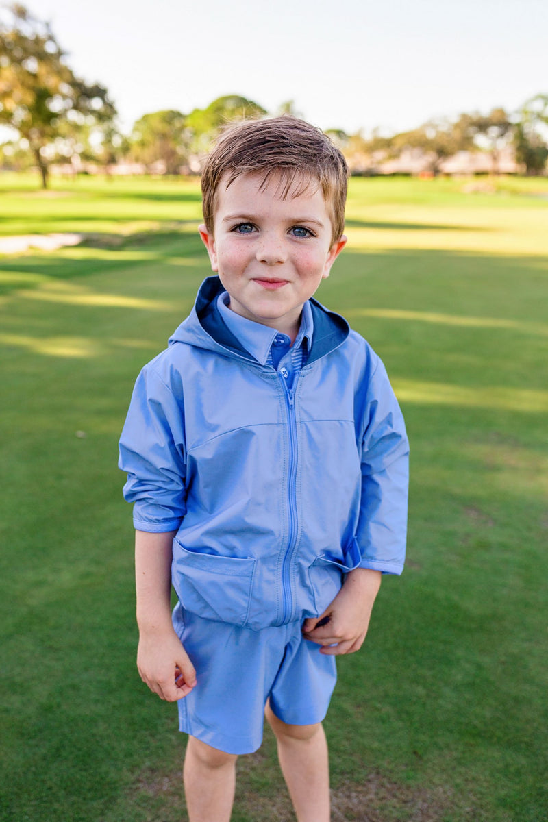 A toddler boy stands on a golf course modeling a light blue zip up hoodie and matching shorts.