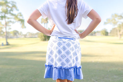 A young girl stands outside on a golf course modeling an outfit. The outfit consists of a white polo shirt and a white golf skort with a blue printed pattern and blue ruffled hem.