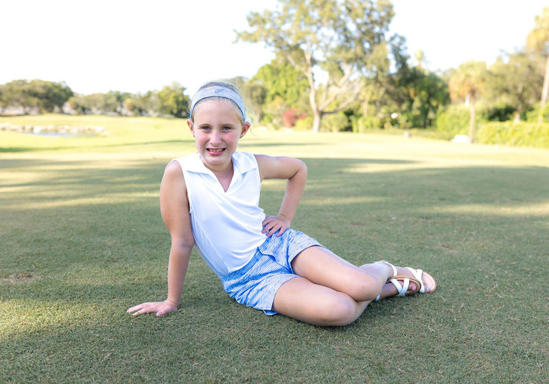 A young girl sits on a golf course modeling a white sleeveless polo shirt and blue chino shorts or blue golf shorts.