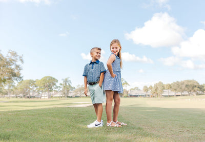 A young boy and girl stand on a golf course modeling golf outfits. The boy is wearing light green chino shorts and a navy blue and green golf polo. The girl is wearing a coordinating blue and white patterned dress.