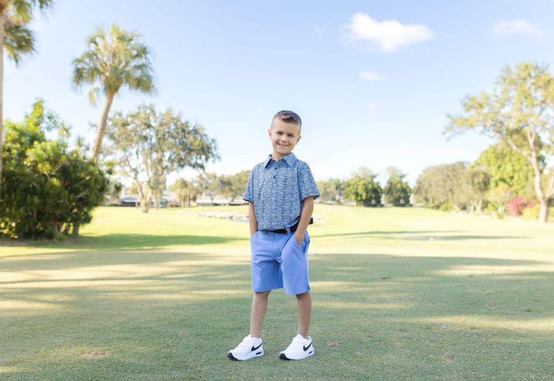 A young boy stands on a golf course modeling a golf outfit consisting of light blue chino shorts and a blue and white stripe patterned polo shirt.