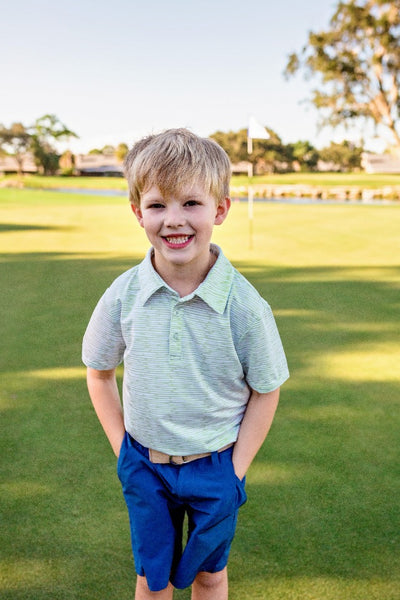 A young boy stands on a golf course modeling navy blue chino shorts and a light green and white striped golf or performance polo shirt.
